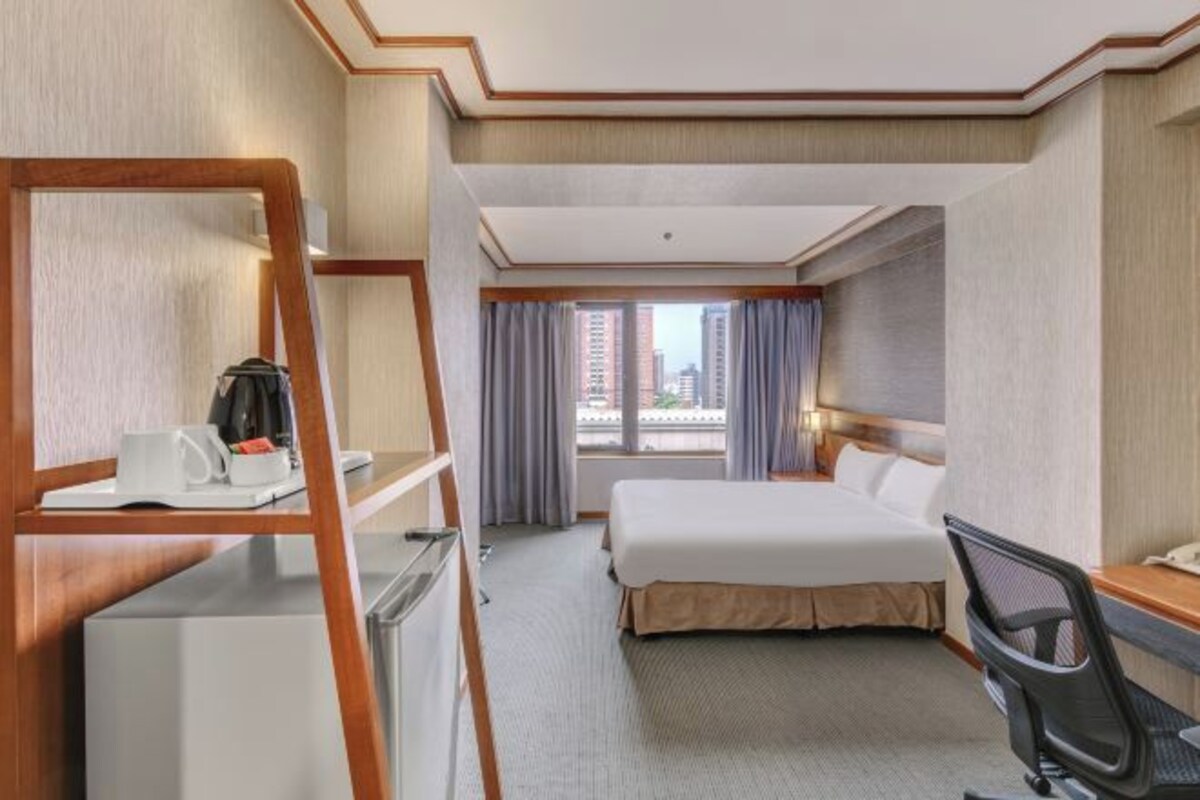 A hotel room with a bed, desk, chair, and a small kitchenette area.