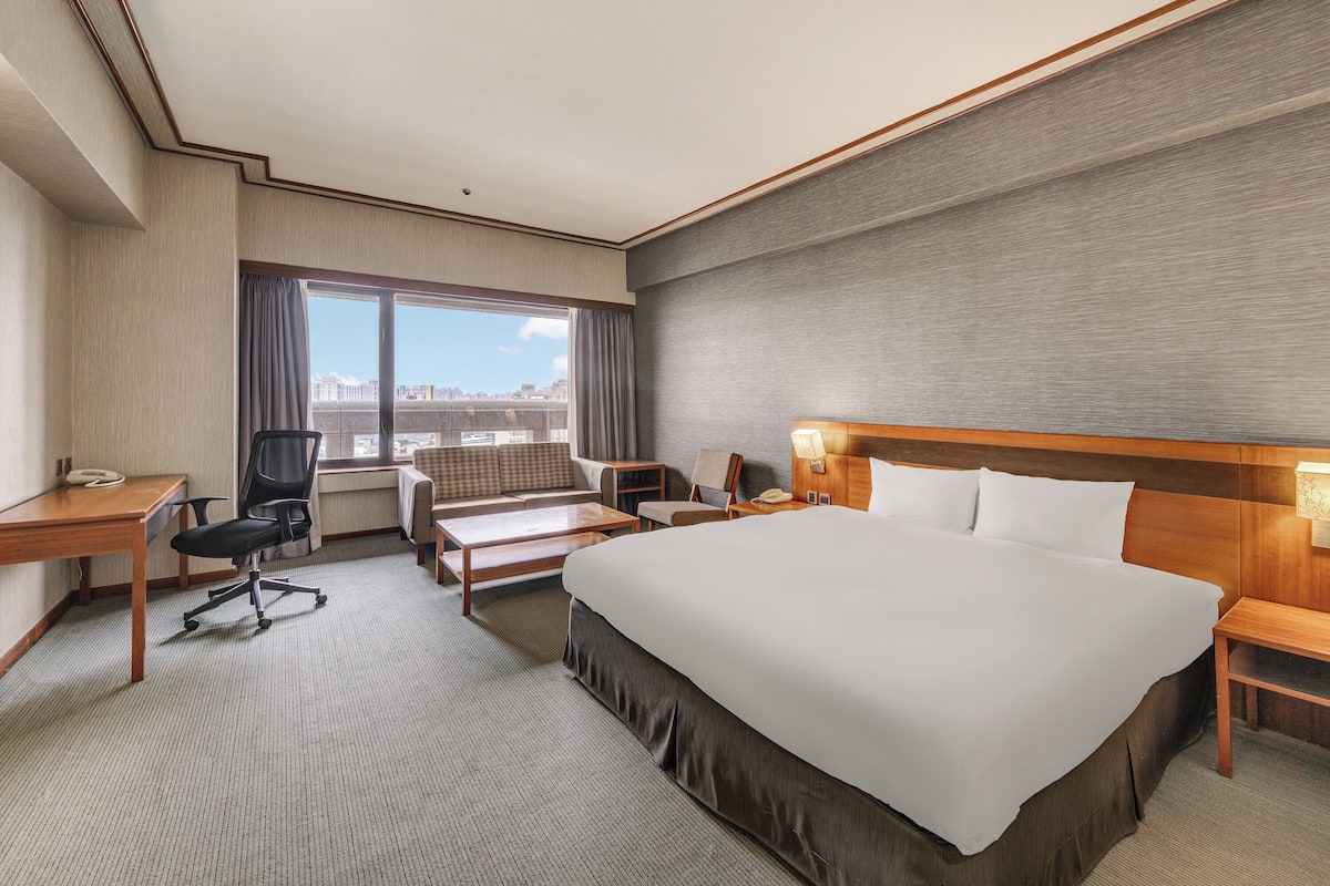 A hotel room with a large bed, a desk, a chair, a sofa, and a view of the city through the window.
