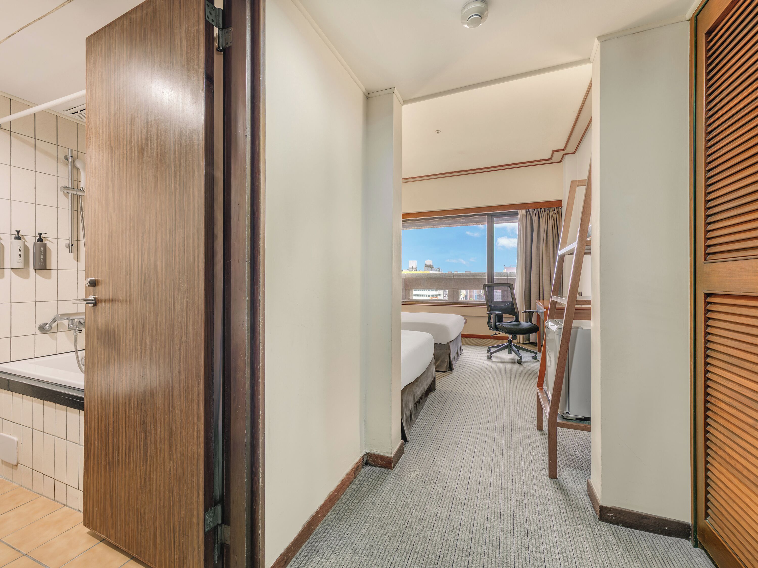 A hotel room with a bathroom, a bed, a desk, and a view of the city.