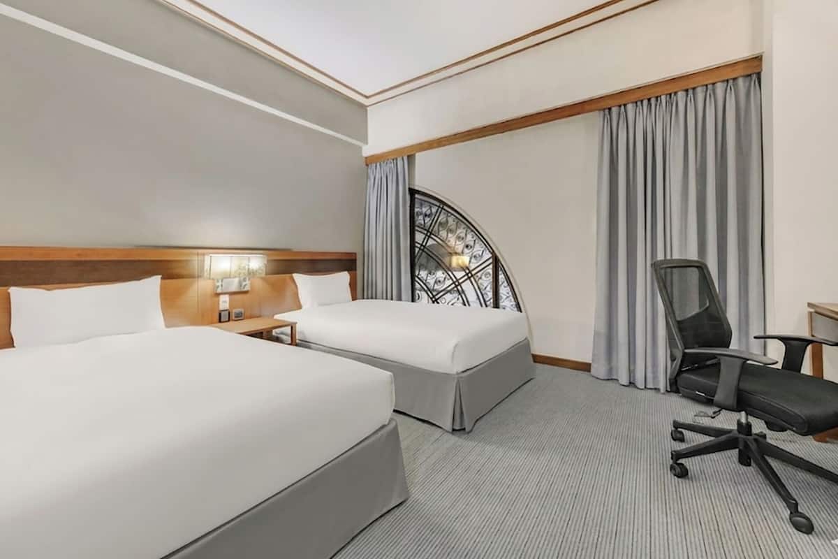 A hotel room with two beds, a desk with a chair, a window with curtains, and a decorative wall piece.