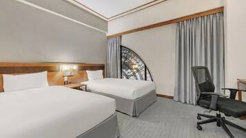 A hotel room with two beds, a desk with a chair, a window with curtains, and a decorative wall piece.
