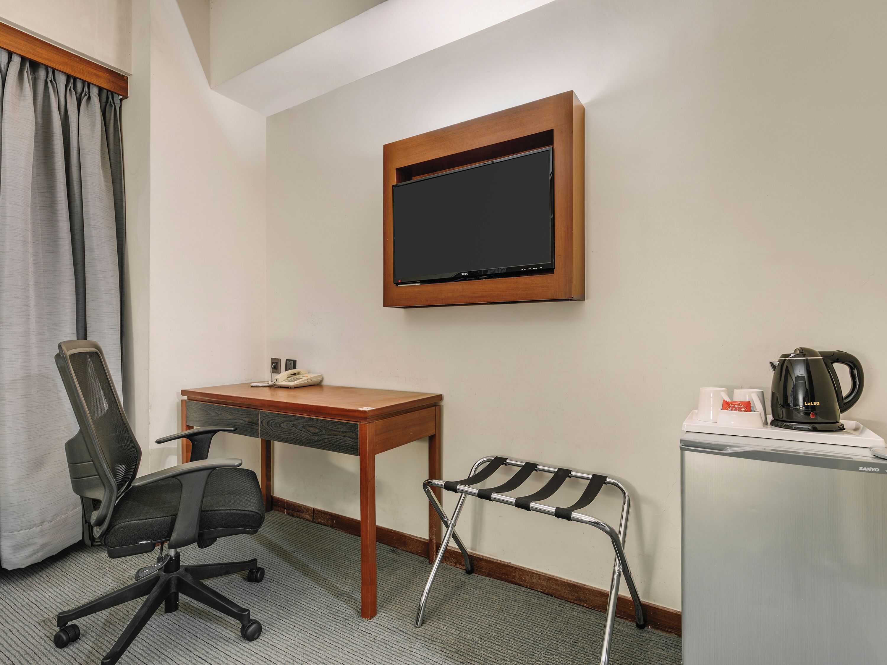 Habitación de hotel con escritorio, silla de oficina, una mesita con hervidor y tazas, una silla alta y televisor fijado en la pared.