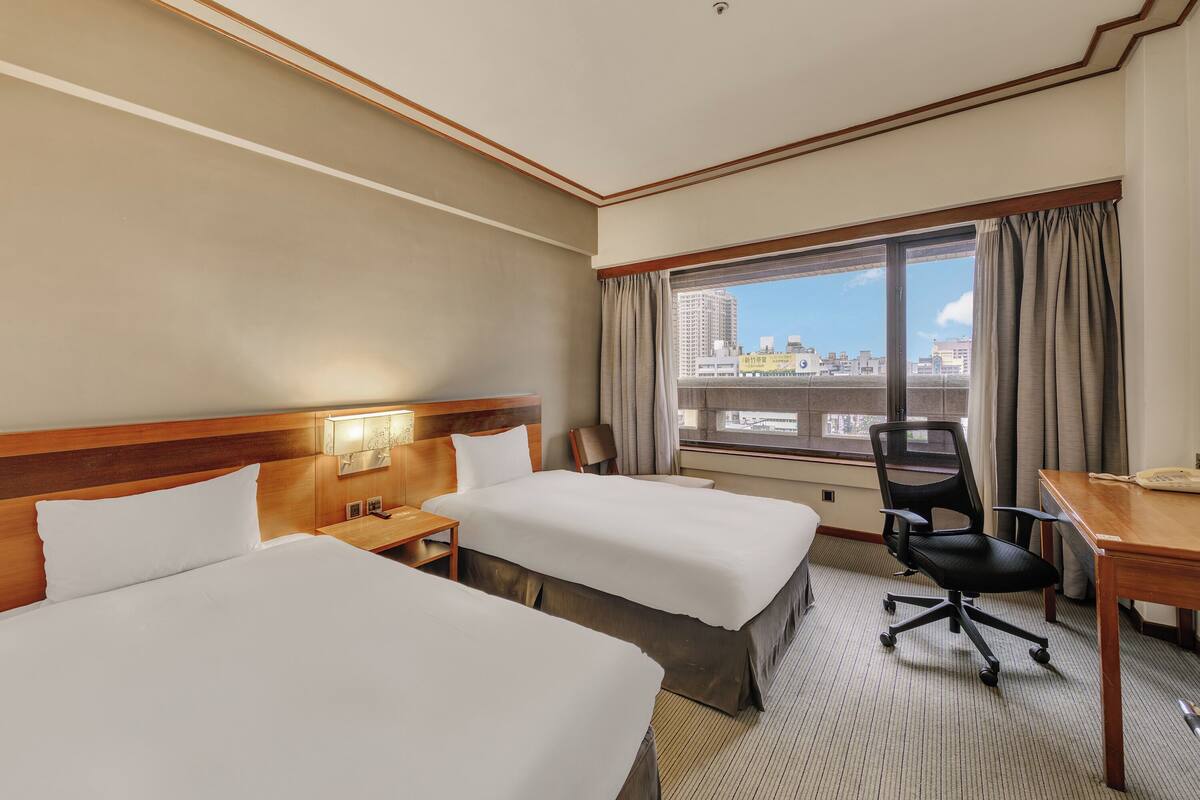 A hotel room with two beds, a desk, a chair, a window with curtains, and a view of the cityscape.