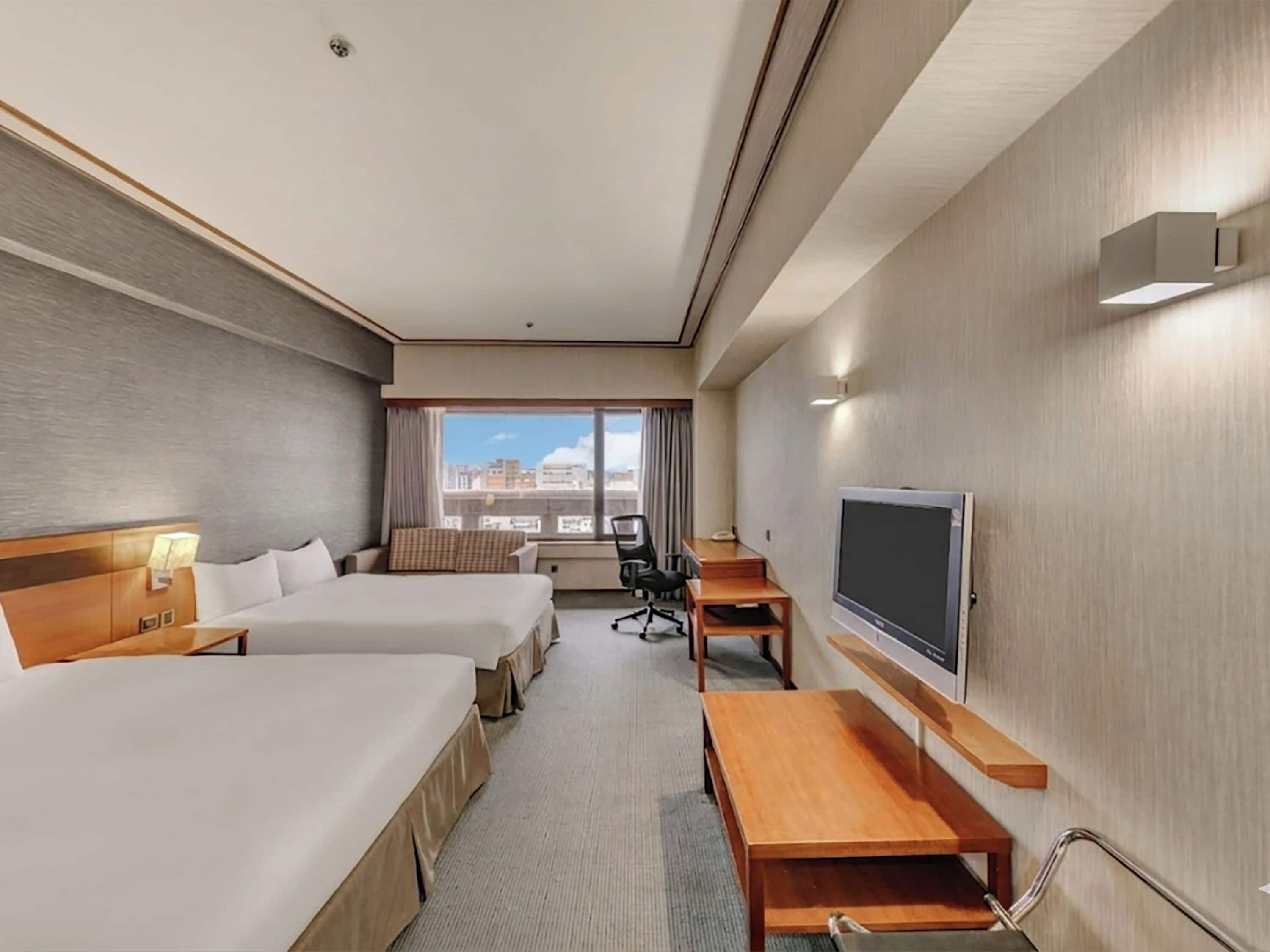 Ein Hotelzimmer mit zwei Betten, einem Schreibtisch, einem Sessel, einem Fernseher und einem Fenster mit Blick auf die Stadt.