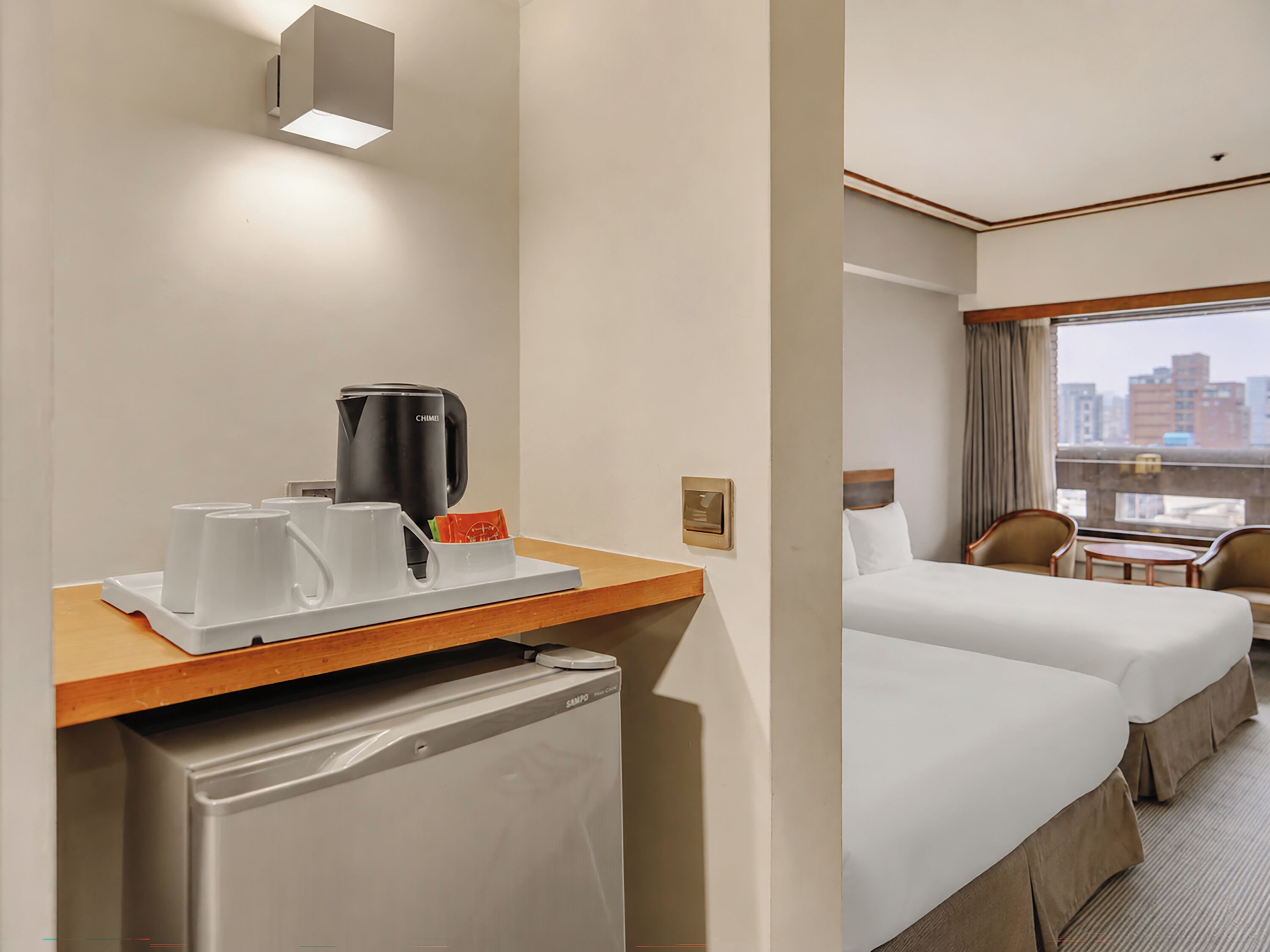 A hotel room with a bed, a desk with a coffee maker, a mini fridge, and a window with a city view.