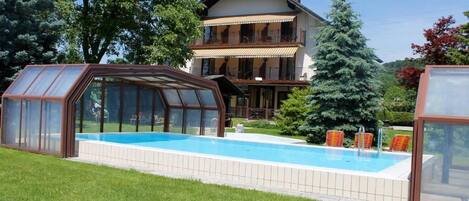 Una casa de dos pisos con piscina, un invernadero de cristal y un gran árbol en el jardín.