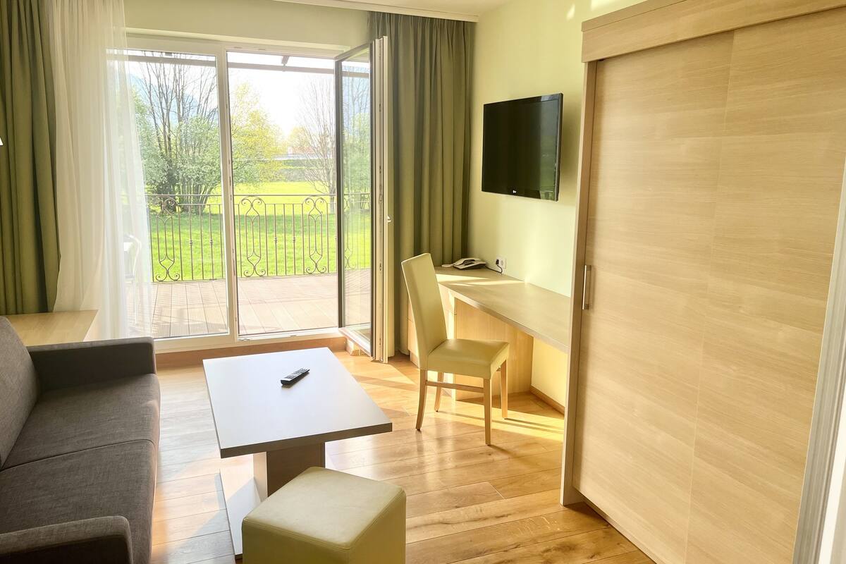 Habitación de hotel con sofá, una mesita, una silla, escritorio, televisor y puerta de cristal corredera que da a un balcón con vistas a zonas verdes.
