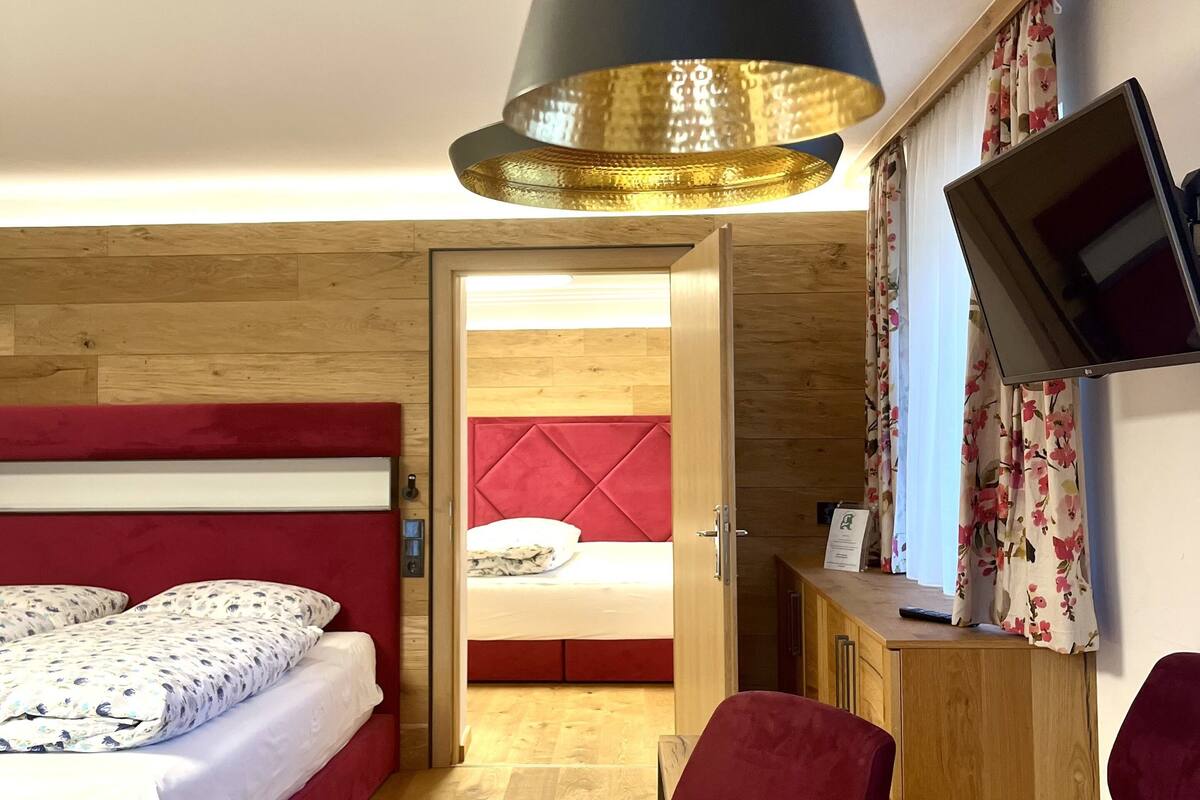 Una habitación de hotel moderna con suelo de madera, una cama tapizada en rojo, un escritorio de madera, un televisor montado en la pared y un amplio ventanal con cortinas florales.
