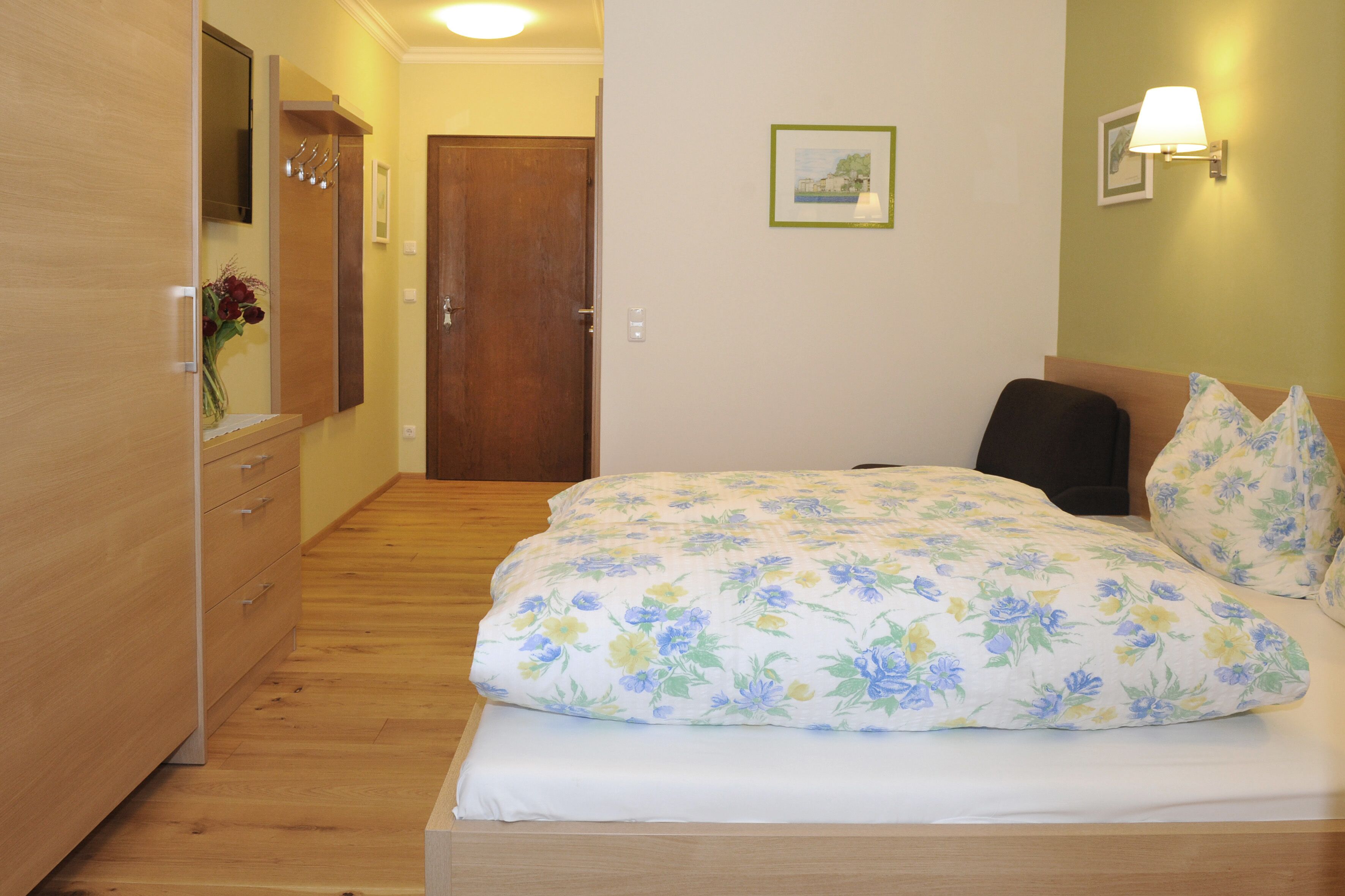 A bedroom with a wooden dresser, a bed with floral bedding, a wooden door, and a wall-mounted lamp.