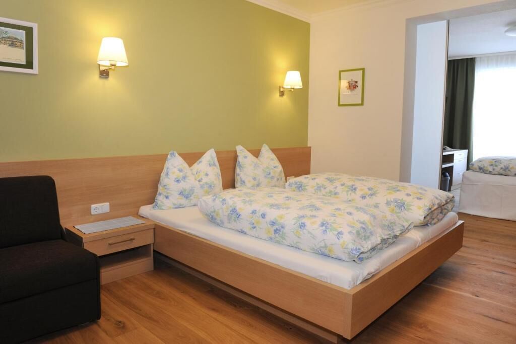 Une chambre d’hôtel avec un lit en bois, une literie à motifs floraux, deux lampes de chevet, une table de chevet, un fauteuil noir et une porte donnant sur une autre pièce.