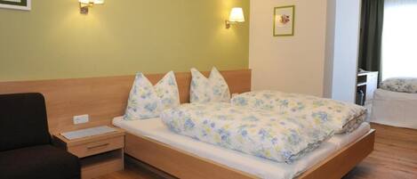 Habitación de hotel con una cama de madera, ropa de cama floral, dos lámparas de noche, una mesita de noche, un sillón negro y una puerta que da a otra habitación.