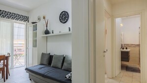 A modern living room with a grey sofa, a dining area with a table and chairs, a clock on the wall, and a view through an open door to a bathroom. - Appartamento Maestrale by Interhome (Viareggio)