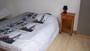 A bedroom with a bed covered in a white duvet, a wooden nightstand with a lamp, and a wall with a light switch.