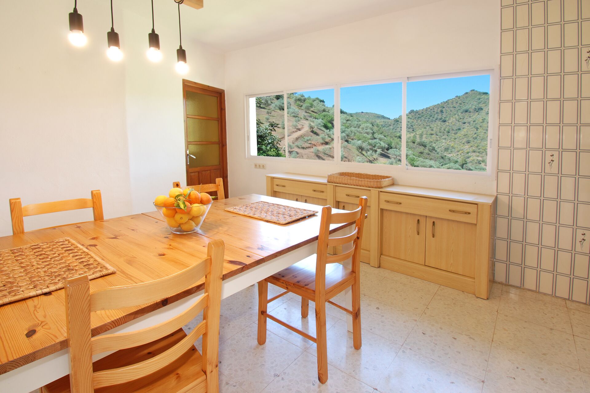 A dining area with a wooden table and chairs, a bowl of fruit, and a view of a hilly landscape through large windows.