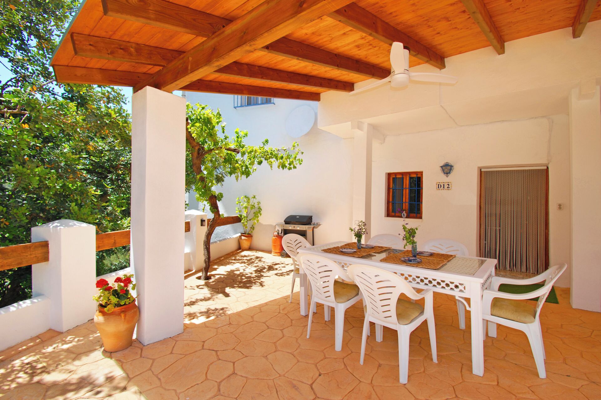 Eine überdachte Terrasse mit Holzboden, einem Tisch mit Stühlen, einem Grill und einem Blumentopf.