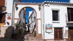 Une rue étroite bordée d’un immeuble blanc, avec une grande porte en arc de cercle, des plantes en pot et une enseigne.