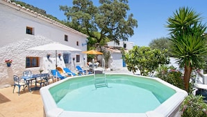 Une piscine entourée de bâtiments blancs et de verdure, avec des chaises longues et des parasols.