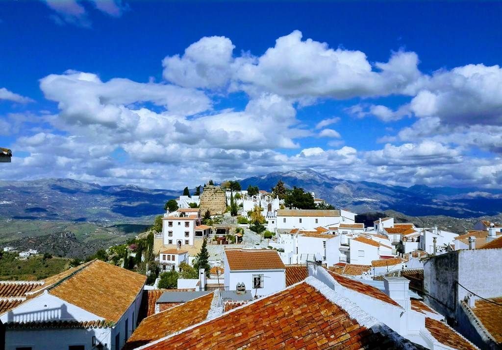 A town with white buildings and red-tiled roofs, set against a backdrop of mountains and a blue sky with clouds.