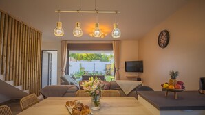 A modern living room with a wooden dining table, a sofa, and a TV. There is a window with a view of greenery outside. - Charming villa between sea and mountains, ideal for friends or family (Saint-Louis)