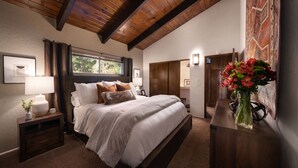 A bedroom with a large bed, wooden headboard, a nightstand with a lamp, a vase with flowers, and a window with curtains.