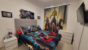 Une chambre à coucher avec un lit agrémenté d’une couvre-lit sur le thème des super-héros, une tête de lit rembourrée de couleur grise, une table de chevet avec une lampe et un téléviseur fixé au mur.