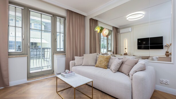 A modern living room with a beige sofa, a white coffee table, and a large window with blinds.
