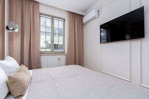 Ein Schlafzimmer mit einem Bett, beigen Vorhängen, einem Fenster mit Jalousien, einem an der Wand montierten Fernseher und einer Klimaanlage.