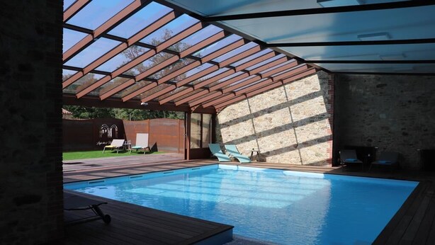 A swimming pool with a wooden deck and a glass ceiling structure. - Gite Aramis (Les Herbiers)