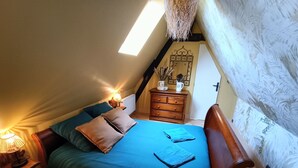 A cozy attic bedroom with a sloped ceiling, a bed with blue bedding, a wooden dresser, and a small table with a lamp. - LONGERE NEAR HONFLEUR, DEAUVILLE, LE HAVRE (Beuzeville)