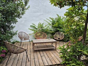 Eine Holzterrasse mit Tisch und drei Korbstühlen, umgeben von üppiger Vegetation und Blick auf ein Gewässer.