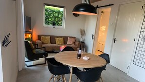 Un comedor con una mesa redonda de madera, sillas negras y una lámpara colgante. Se ve un sofá y una ventana con follaje al fondo.