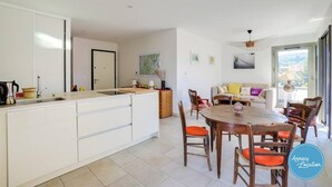Una cocina moderna con armarios blancos, un comedor con sillas de madera y una mesa redonda, y un área de estar con sofá y vistas al exterior.