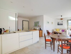 Une cuisine moderne avec des meubles blancs, un coin repas avec des chaises en bois et une table ronde, et un salon avec un canapé et une vue sur l’extérieur.