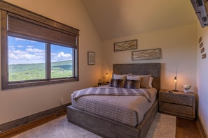 A bedroom with a large window, a wooden headboard, a bedside table with a lamp, and a view of a green landscape.