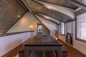 A ping pong table in a room with wooden beams, a billiards rack, and a window with blinds.