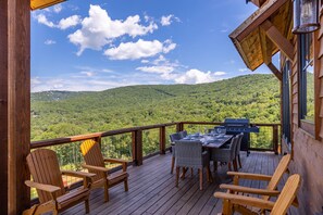 A wooden deck with a table and chairs, overlooking a forested mountain range.