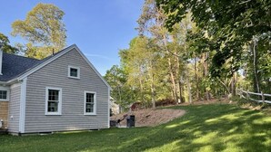 A two-story house with a sloped roof, a front yard with a wooden fence, and a wooded area in the background. - New construction within 1 mile of bay beach and Nickerson Park (Brewster)