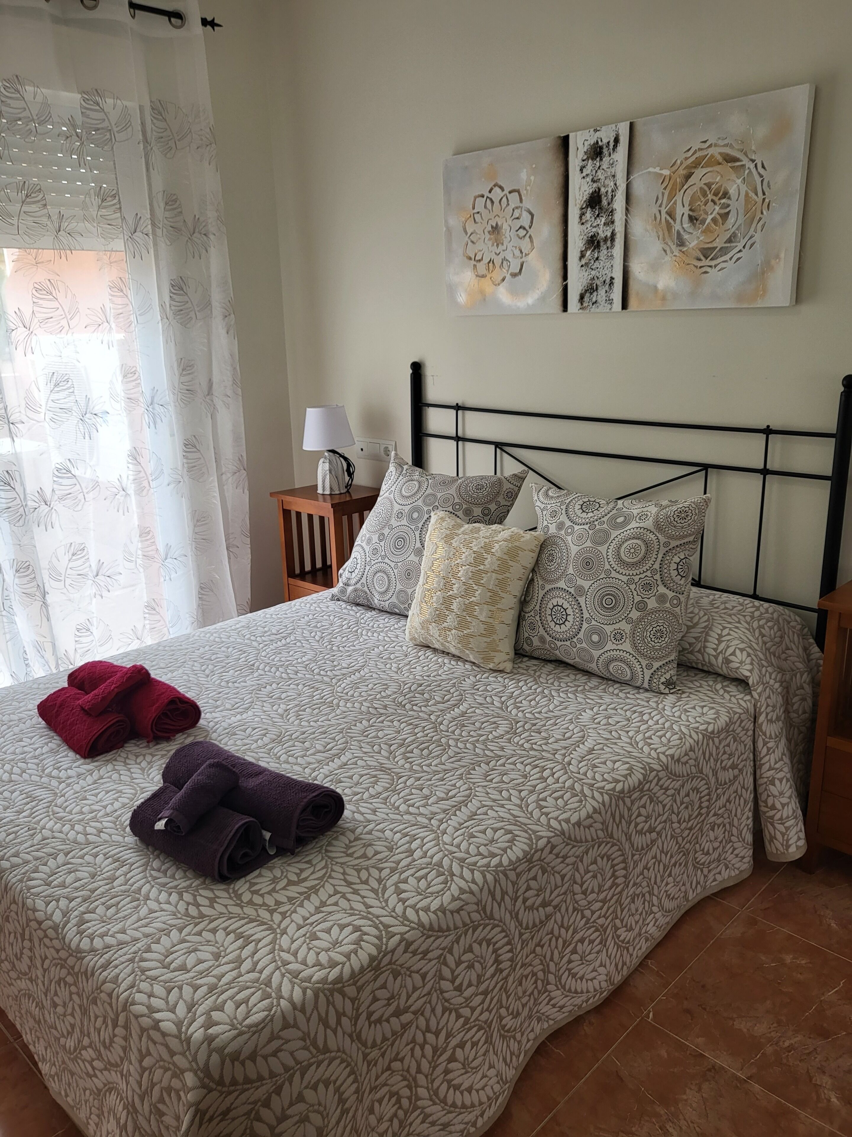 A neatly made bed with patterned bedding, two rolled towels, and a bedside table with a lamp.