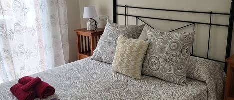 A neatly made bed with patterned bedding, two rolled towels, and a bedside table with a lamp.