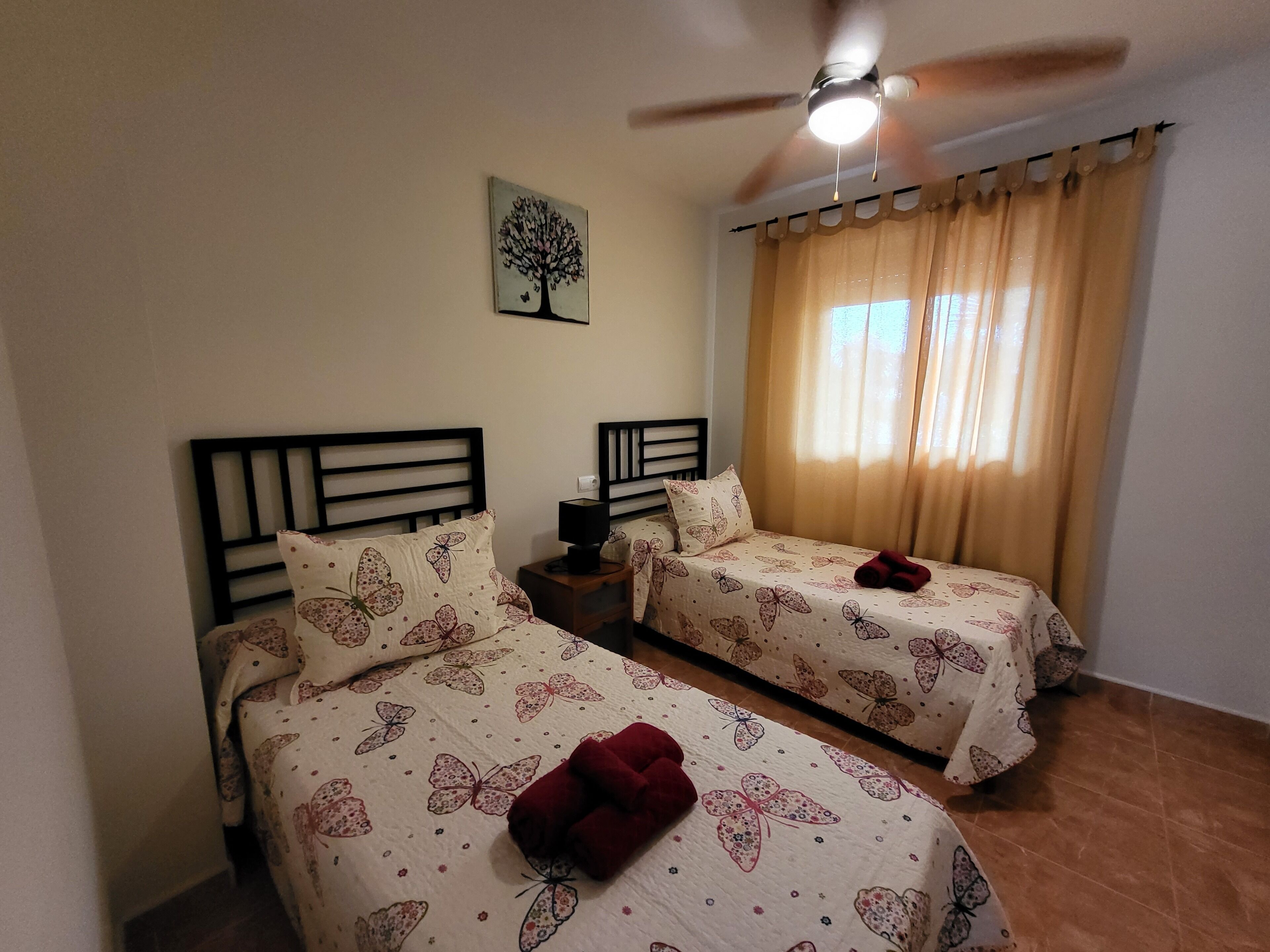 A hotel room with two single beds, a ceiling fan, a nightstand with a lamp, and a window with curtains.