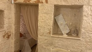 Una habitación rústica con pared de piedra, una mesa de madera con un plato, una silla blanca y un pequeño estante con un candelabro.
