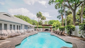 Une piscine dotée de chaises longues et d’un espace de repos couvert, le tout entouré d’arbres et d’un bâtiment.