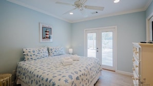 Une chambre à coucher comprenant un lit, un ventilateur de plafond, un tableau encadré, une commode et une porte donnant sur l’extérieur.