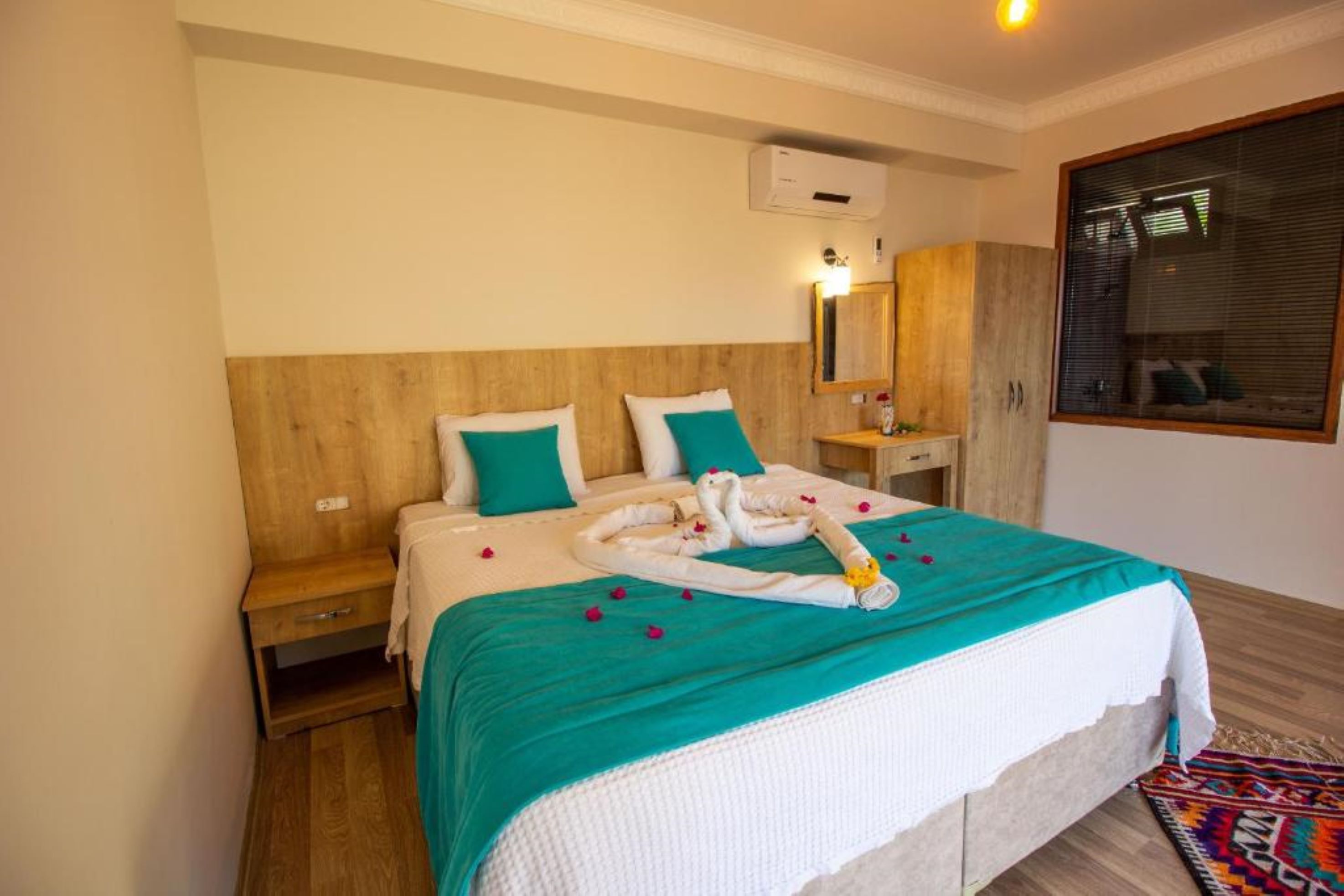 Une chambre d’hôtel avec une tête de lit en bois, un lit avec une literie de couleur turquoise, une table de chevet, un miroir et une fenêtre avec des stores.