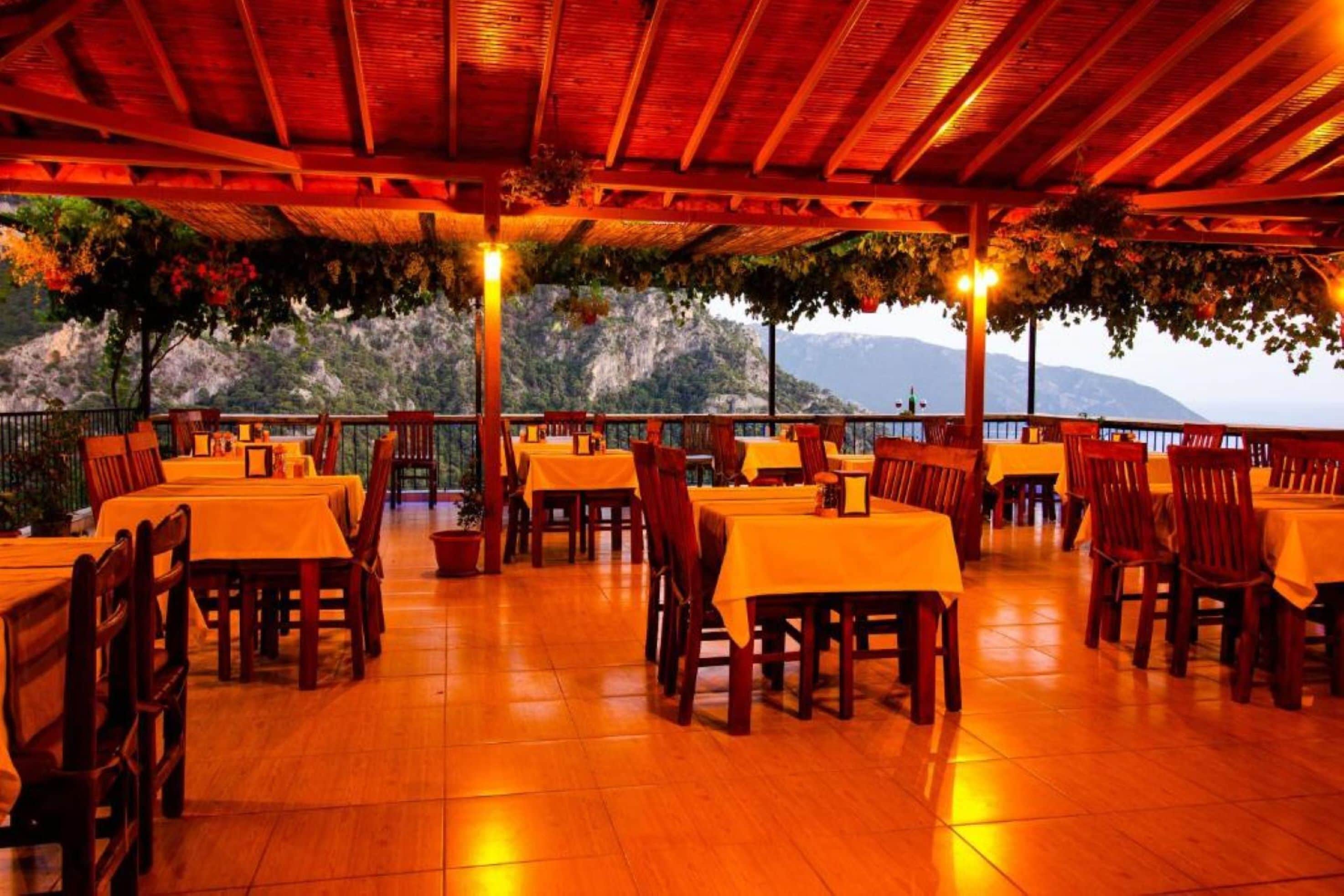 An outdoor dining area with tables and chairs, a wooden ceiling, and a view of a mountainous landscape.
