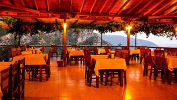 An outdoor dining area with tables and chairs, a wooden ceiling, and a view of a mountainous landscape.