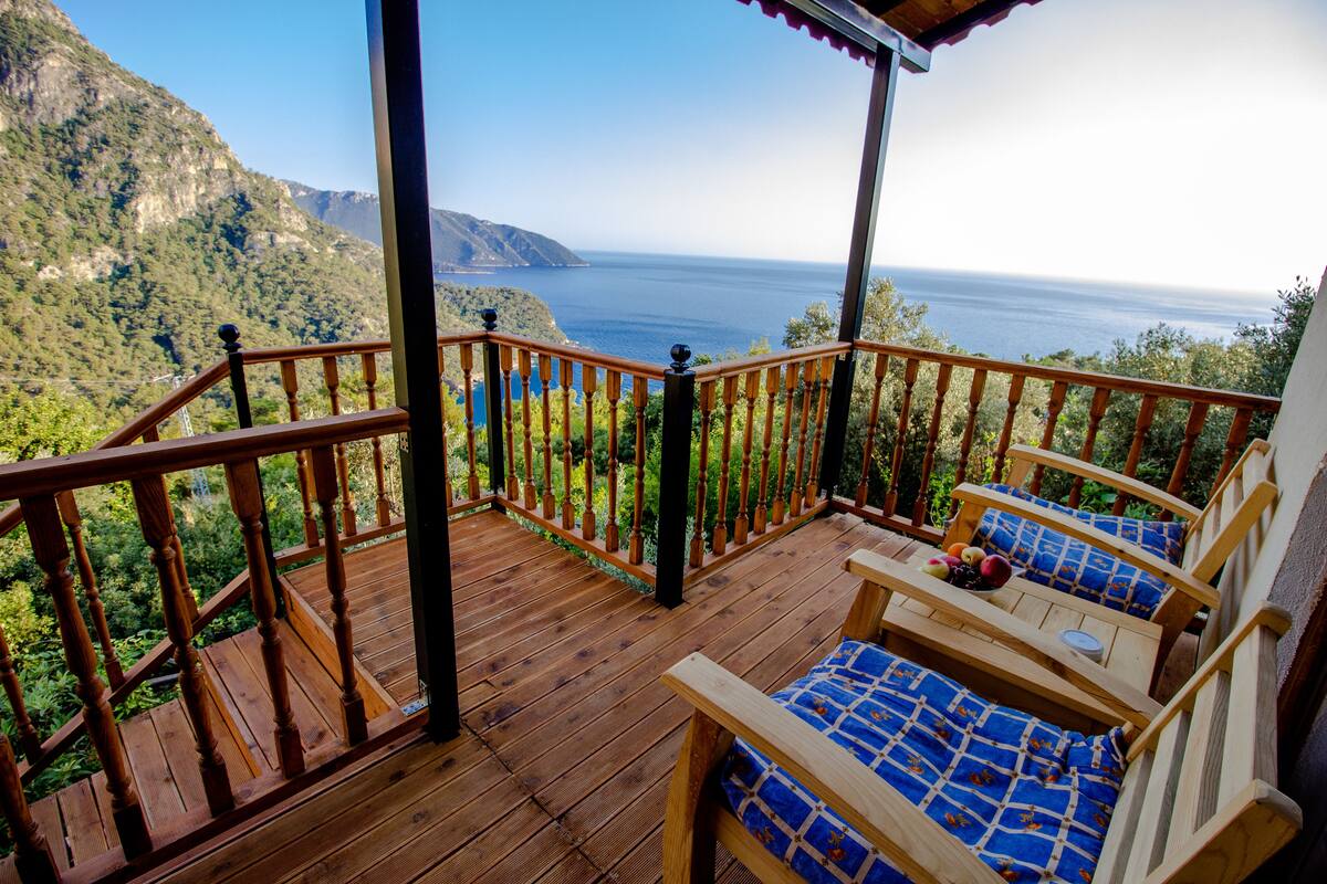 A wooden deck with a view of the ocean and mountains. There are two wooden chairs with blue cushions.