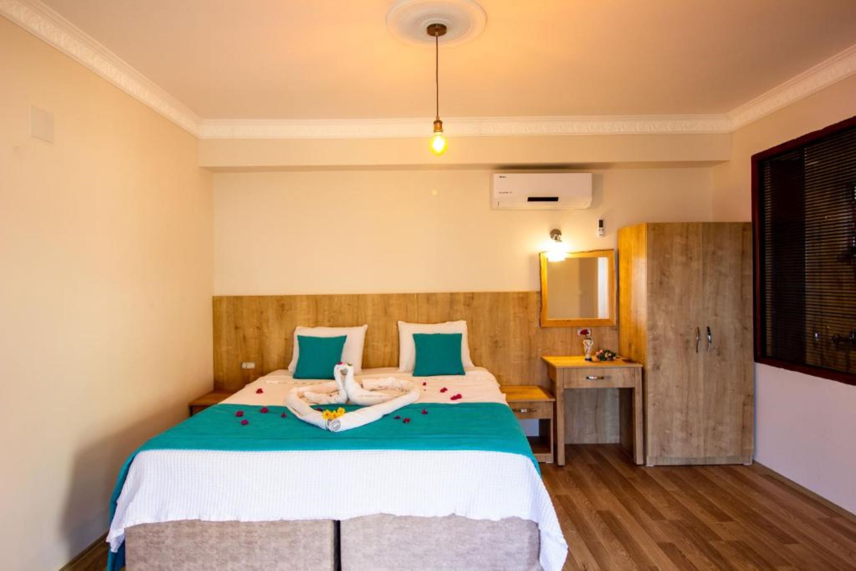 Een hotelkamer met een houten hoofdbord, een bed met een blauwgroene sprei, een nachtkastje met een lamp, een houten garderobe en een raam met jaloezieën.