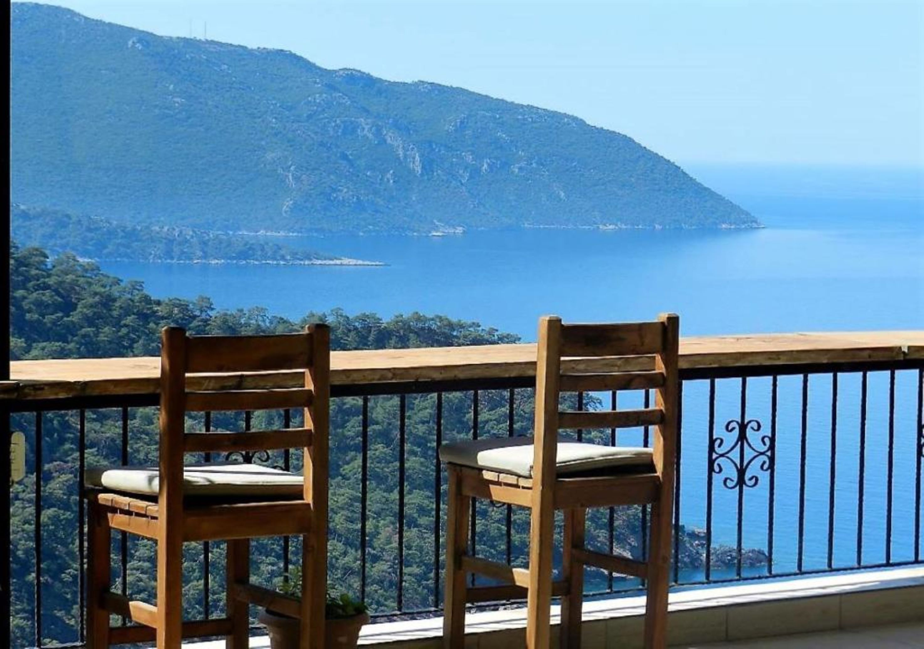 Two wooden chairs with cushions on a balcony overlooking a mountainous coastline.