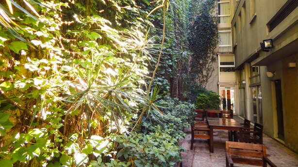 Un callejón estrecho con abundante vegetación a un lado y una fila de mesas y sillas de madera al otro.